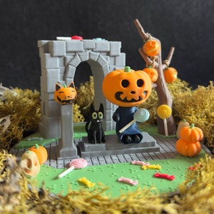 May include: A miniature Halloween scene featuring a pumpkin-headed figure holding a lollipop, a black cat, and a stone archway. The scene is decorated with small pumpkins, sweets, and moss.