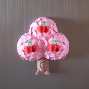 May include: A whimsical pink tree-shaped decoration with three clusters of red cherries. The tree has a textured, layered design with a light brown trunk. The cherries have green stems. The background is a neutral gray.