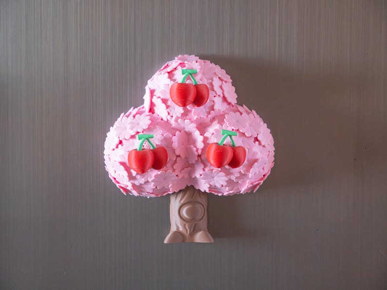 May include: A whimsical pink tree-shaped decoration with three clusters of red cherries. The tree has a textured, layered design with a light brown trunk. The cherries have green stems. The background is a neutral gray.