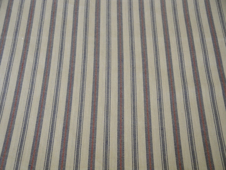 May include: Cream-coloured fabric with vertical stripes. Alternating stripes of navy blue and thin red lines create a classic pattern. The fabric has a textured appearance, suggesting a woven material.