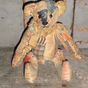 May include: A vintage teddy bear made from patchwork fabric. The bear has a tan body with patches of blue, red, and patterned fabric. It has black button eyes and a tan scarf around its neck. The bear is sitting on a wooden surface.