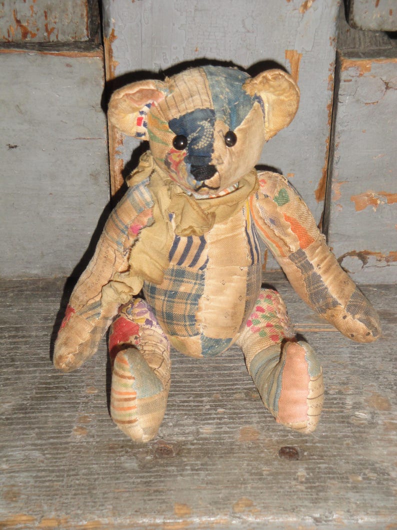 May include: A vintage teddy bear made from patchwork fabric. The bear has a tan body with patches of blue, red, and patterned fabric. It has black button eyes and a tan scarf around its neck. The bear is sitting on a wooden surface.