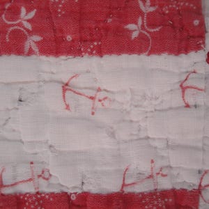 Op de afbeelding: Een gewatteerde textielstof met afwisselende horizontale banden in rood en wit. De rode banden tonen een wit bloemmotief, terwijl de witte banden zijn versierd met een herhalend rood ankermotief. De stof heeft een gekreukte textuur.