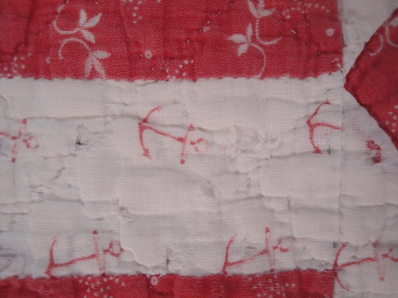 Op de afbeelding: Een gewatteerde textielstof met afwisselende horizontale banden in rood en wit. De rode banden tonen een wit bloemmotief, terwijl de witte banden zijn versierd met een herhalend rood ankermotief. De stof heeft een gekreukte textuur.