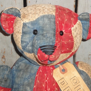 May include: A patchwork quilt teddy bear with blue, red, and white fabric. The bear has black button eyes and a black embroidered nose. The bear is holding a brown tag with the text "Old Blue" in block letters.
