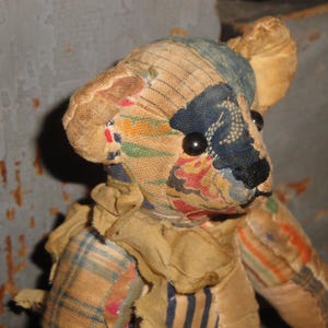 May include: A vintage teddy bear with a patchwork design. The bear's head features a blue and floral patterned fabric, with black button eyes and a black nose. The bear's body is made of various fabrics, including plaid and striped patterns.