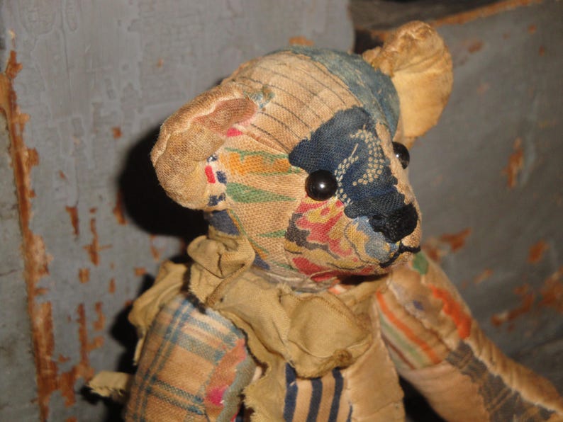 May include: A vintage teddy bear with a patchwork design. The bear's head features a blue and floral patterned fabric, with black button eyes and a black nose. The bear's body is made of various fabrics, including plaid and striped patterns.
