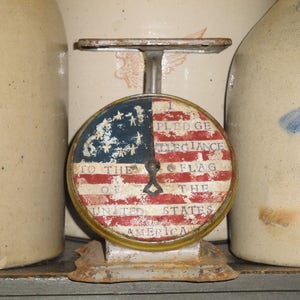 May include: A vintage scale with a round face featuring an American flag design. The face reads "I pledge allegiance to the flag of the United States of America".