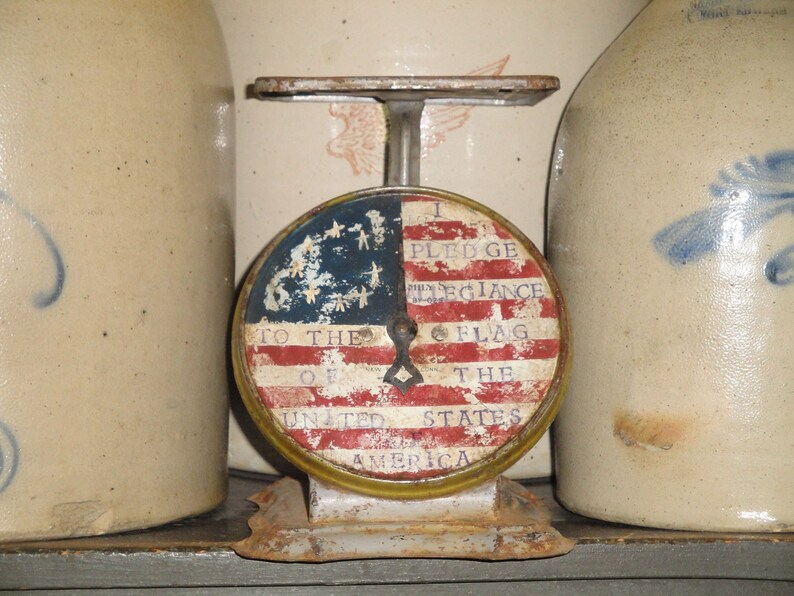 May include: A vintage scale with a round face featuring an American flag design. The face reads "I pledge allegiance to the flag of the United States of America".