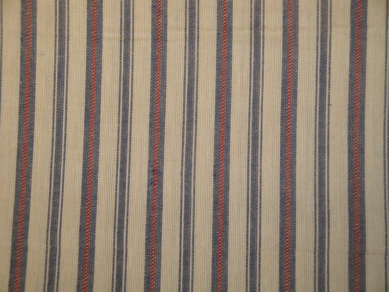 May include: Cream-coloured fabric with vertical stripes. Alternating stripes of navy blue and red run the length of the fabric. The fabric has a woven texture, and the stripes are evenly spaced.