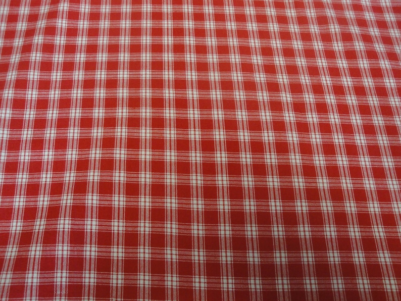 May include: Close-up of a red and white plaid fabric. The pattern consists of a grid of white lines intersecting over a red background, creating a classic checkered design. The fabric appears to be cotton or a similar woven material.