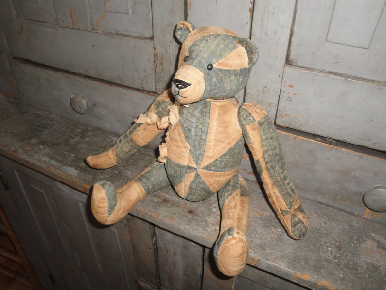 May include: A vintage patchwork teddy bear with a blue and tan color scheme. The bear is sitting on a wooden surface.