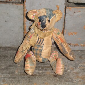May include: A vintage teddy bear with a patchwork design. The bear has a tan body with patches of blue, plaid, and floral fabrics. It has black button eyes and a stitched nose. The bear is sitting on a wooden surface.