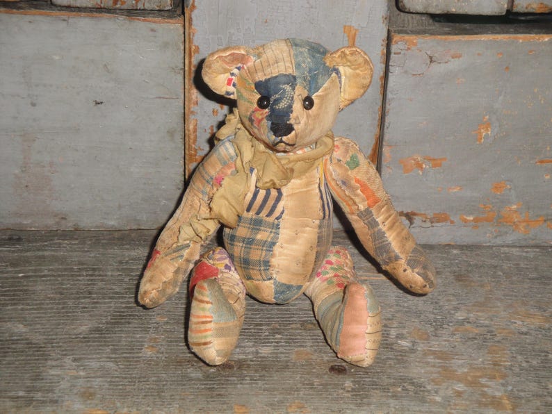 May include: A vintage teddy bear with a patchwork design. The bear has a tan body with patches of blue, plaid, and floral fabrics. It has black button eyes and a stitched nose. The bear is sitting on a wooden surface.