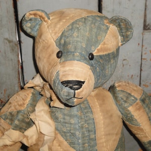 May include: A patchwork teddy bear with a blue and tan fabric design. The bear has black button eyes and a black stitched nose.