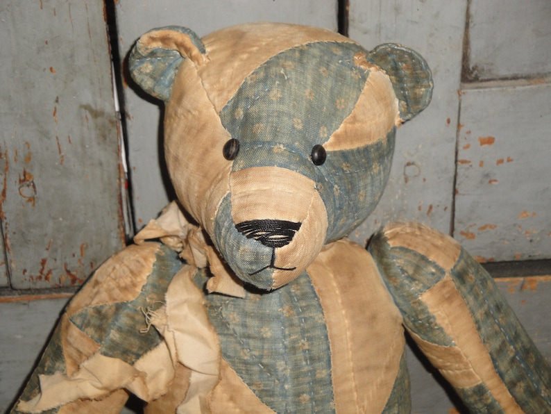 May include: A patchwork teddy bear with a blue and tan fabric design. The bear has black button eyes and a black stitched nose.