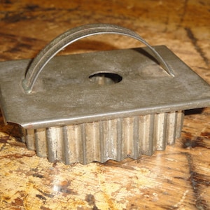 May include: A vintage, square-shaped cookie cutter with a corrugated edge and a curved handle. The metal cutter has a worn, antique appearance, suggesting it has been used for baking. The handle is arched for easy use.
