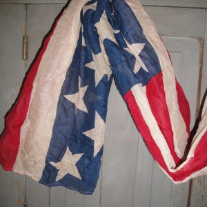 Op de afbeelding: Een vintage Amerikaanse vlag sjaal in rood, wit en blauw met een vervaagde look. De sjaal is gedrapeerd over een houten deur.