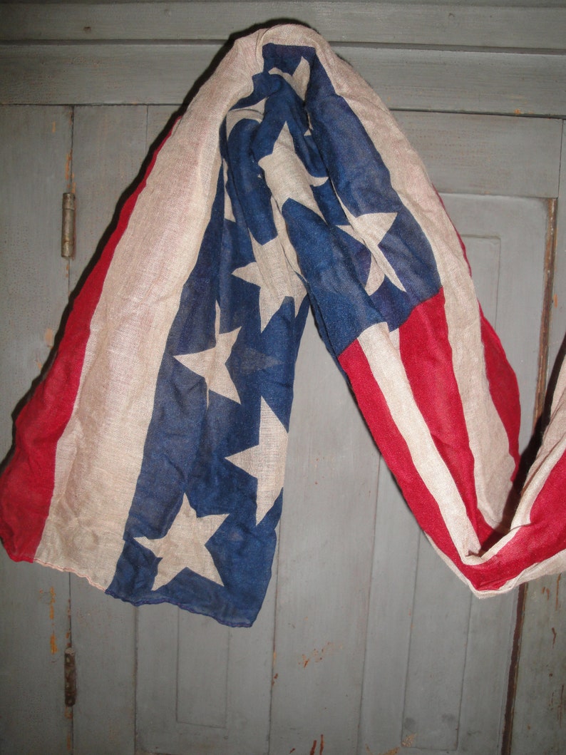 Op de afbeelding: Een vintage Amerikaanse vlag sjaal in rood, wit en blauw met een vervaagde look. De sjaal is gedrapeerd over een houten deur.