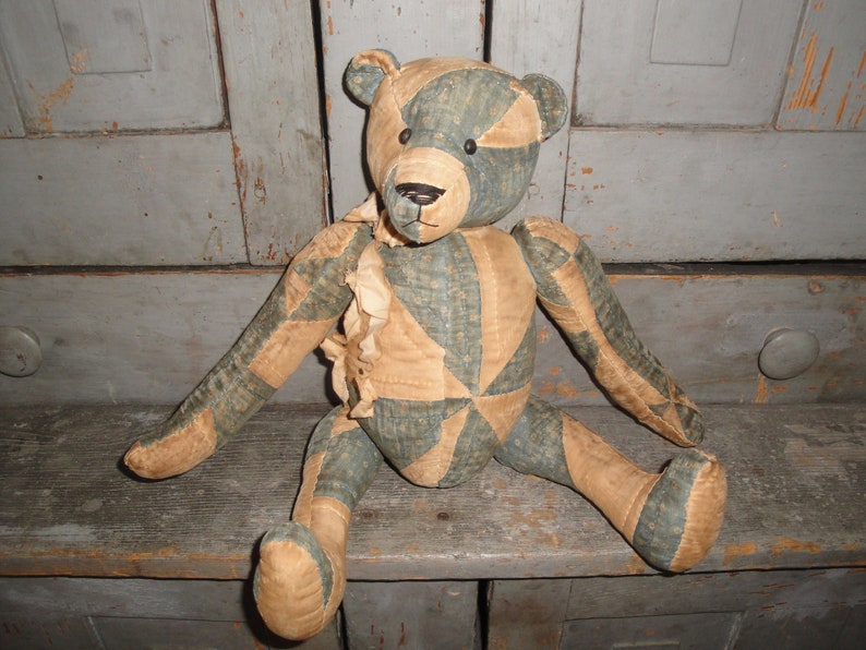 May include: A vintage patchwork teddy bear with a blue and tan patterned fabric. The bear is sitting on a wooden surface.