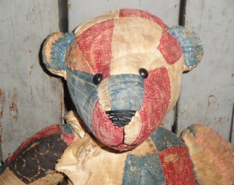 May include: A vintage patchwork teddy bear with a stitched black nose and button eyes. The bear's head is composed of red, blue, and cream-colored fabric patches. The bear's ears are blue and cream-colored.