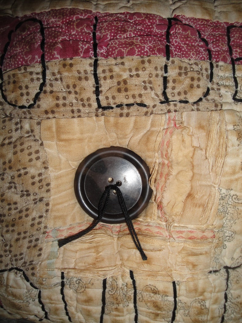 May include: A close-up of a vintage quilt with a brown button and black thread. The quilt is made of different fabrics and has a patchwork design. The word "OLD" is stitched in black thread on the quilt.