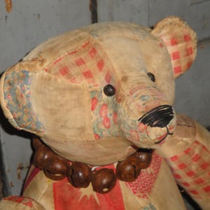 May include: A vintage patchwork teddy bear with a red and white checkered pattern on one ear. The bear has a brown bead necklace and a black embroidered nose.