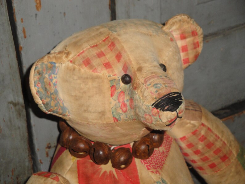 May include: A vintage patchwork teddy bear with a red and white checkered pattern on one ear. The bear has a brown bead necklace and a black embroidered nose.