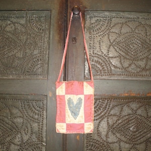 Op de afbeelding: Een kleine, handgemaakte schoudertas met een patchwork-ontwerp. De tas heeft een roze en cr&egrave;mekleurig geblokt patroon met een grijze hart in het midden. De tas hangt aan een dunne, roze band tegen een getextureerde, donkergroene achtergrond.