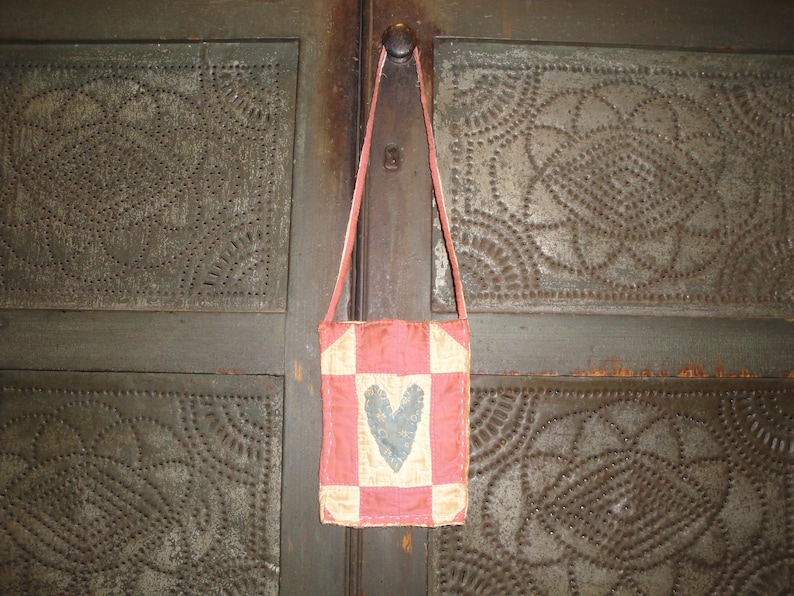 Op de afbeelding: Een kleine, handgemaakte schoudertas met een patchwork-ontwerp. De tas heeft een roze en cr&egrave;mekleurig geblokt patroon met een grijze hart in het midden. De tas hangt aan een dunne, roze band tegen een getextureerde, donkergroene achtergrond.