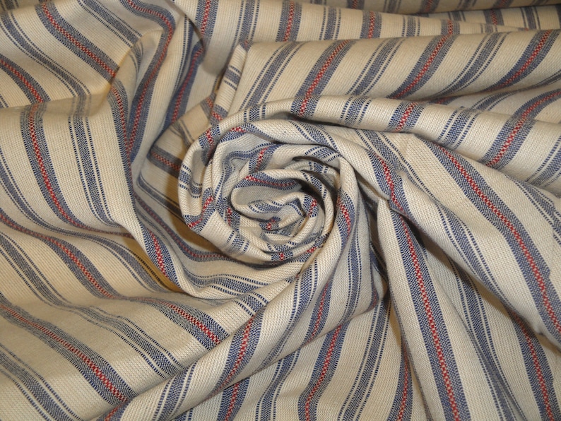 May include: A close-up of a fabric with a cream background and vertical stripes. The stripes alternate between blue and red, creating a classic, textured pattern. The fabric is gathered and folded, showing the texture and drape of the material.