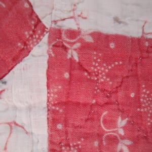 Op de afbeelding: Een gewatteerd textiel met een patchworkontwerp van afwisselende witte en rode vierkanten. De rode secties zijn versierd met een wit bloemen- en stippenpatroon, terwijl de witte secties vage rode lijntekeningen vertonen.
