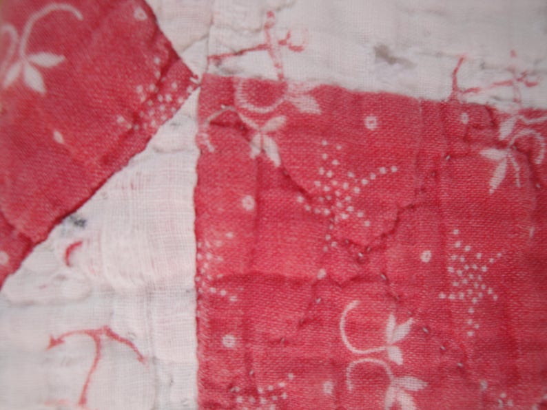 Op de afbeelding: Een gewatteerd textiel met een patchworkontwerp van afwisselende witte en rode vierkanten. De rode secties zijn versierd met een wit bloemen- en stippenpatroon, terwijl de witte secties vage rode lijntekeningen vertonen.