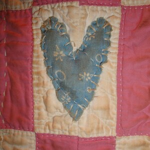 Op de afbeelding: Close-up van een handgemaakte quilt met een blauw hart applicatie. De quilt heeft een patchwork ontwerp met afwisselende vierkanten van roze en gebroken witte stof. Het hart is gestikt met een decoratieve rand.