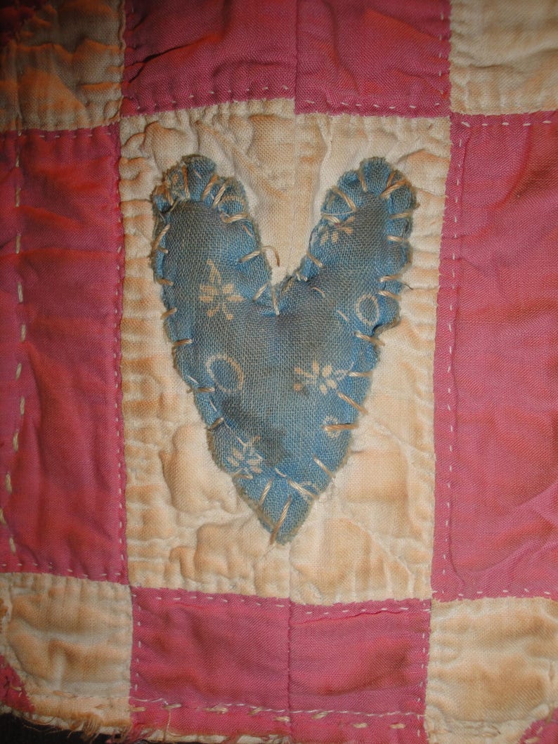 Op de afbeelding: Close-up van een handgemaakte quilt met een blauw hart applicatie. De quilt heeft een patchwork ontwerp met afwisselende vierkanten van roze en gebroken witte stof. Het hart is gestikt met een decoratieve rand.