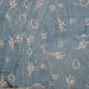May include: A light blue fabric featuring a white floral and circular pattern with visible white stitching lines running across the surface. The material is composed of multiple overlapping pieces of textile joined together in a patchwork style.