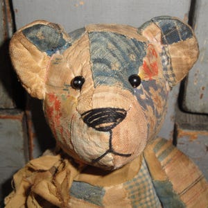 Puede incluir: Un oso de peluche construido con varios parches de tela en tonos azul, bronceado y naranja descolorido. El oso presenta dos peque&ntilde;os ojos de cuentas negras, una nariz y boca cosidas a mano con hilo negro y una bufanda de tela bronceada deshilachada.