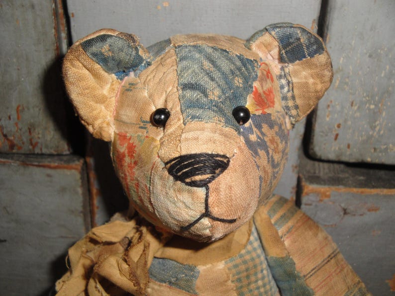 Puede incluir: Un oso de peluche construido con varios parches de tela en tonos azul, bronceado y naranja descolorido. El oso presenta dos peque&ntilde;os ojos de cuentas negras, una nariz y boca cosidas a mano con hilo negro y una bufanda de tela bronceada deshilachada.