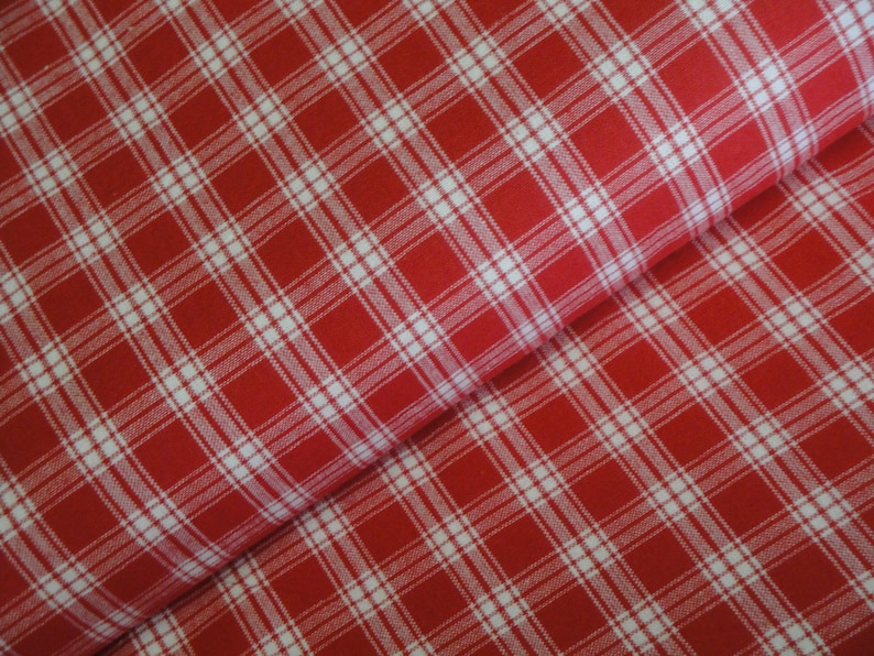 May include: Close-up of red and white plaid fabric. The fabric features a classic plaid pattern with intersecting white lines on a red background. The fabric is folded, showing the texture and drape of the material.