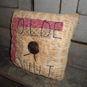 May include: A cream-colored pillow with a red and white patchwork design. The pillow is embroidered with the words "OLDE QUILT" in black thread. A brown button is sewn onto the pillow.