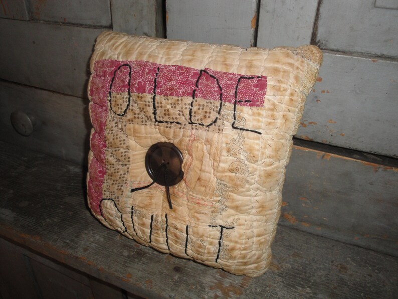 May include: A cream-colored pillow with a red and white patchwork design. The pillow is embroidered with the words "OLDE QUILT" in black thread. A brown button is sewn onto the pillow.