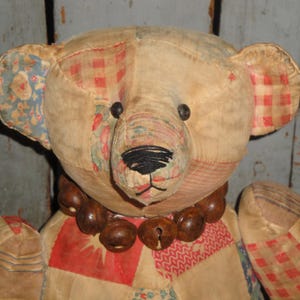 May include: A vintage patchwork teddy bear with a brown bead necklace. The bear has a light brown fur and is made of different fabrics, including red and white gingham, blue and white floral, and a cream-colored fabric with a geometric pattern.