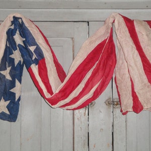 Op de afbeelding: Een vintage Amerikaanse vlag gedrapeerd over een houten deur. De vlag is verbleekt en versleten, met rode, witte en blauwe strepen en een veld met blauwe sterren.
