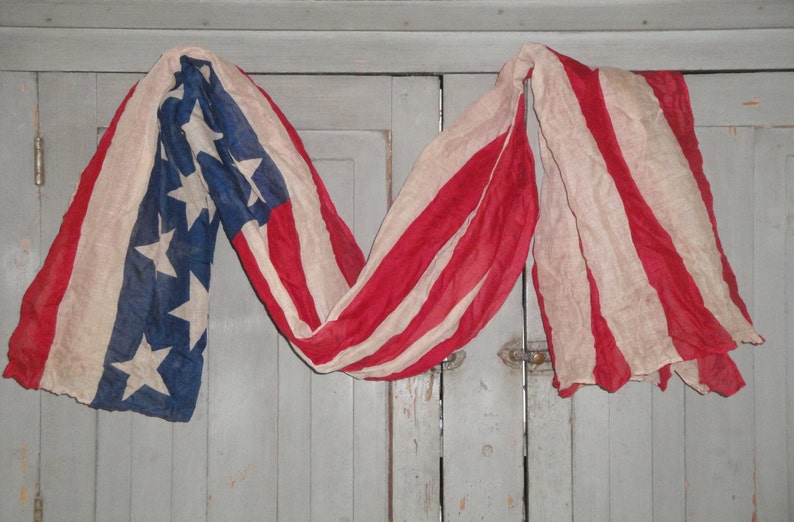 Op de afbeelding: Een vintage Amerikaanse vlag gedrapeerd over een houten deur. De vlag is verbleekt en versleten, met rode, witte en blauwe strepen en een veld met blauwe sterren.