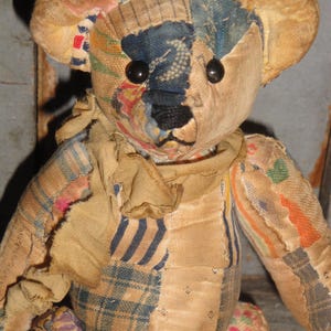 May include: A vintage teddy bear with a patchwork design. The bear features a mix of fabrics in various colors, including blue, beige, and red. It has black button eyes and a stitched nose. The bear's arms and legs are attached, and it has a scarf around its neck.