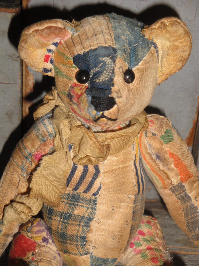May include: A vintage teddy bear with a patchwork design. The bear features a mix of fabrics in various colors, including blue, beige, and red. It has black button eyes and a stitched nose. The bear's arms and legs are attached, and it has a scarf around its neck.