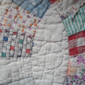 May include: A patchwork quilt featuring a variety of fabric squares in white, red, blue, and green. The design includes floral patterns, checkered prints, and striped textures, all stitched together with visible quilting lines on a white fabric base.