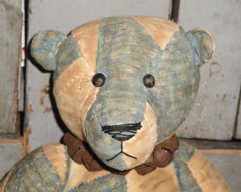 May include: A patchwork teddy bear with blue and tan fabric. The bear has black button eyes and a black stitched nose. The bear is wearing a brown fabric collar with bells.