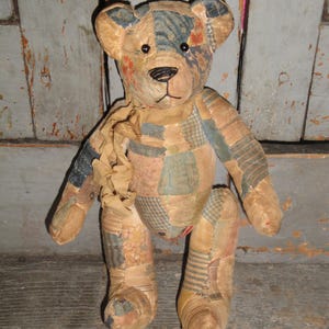 Puede incluir: Un oso de peluche confeccionado con varios parches de tela en tonos apagados de azul, beige y crema. El oso presenta ojos redondos oscuros, una nariz y boca negras bordadas, y una cinta de tela fruncida atada alrededor del cuello.