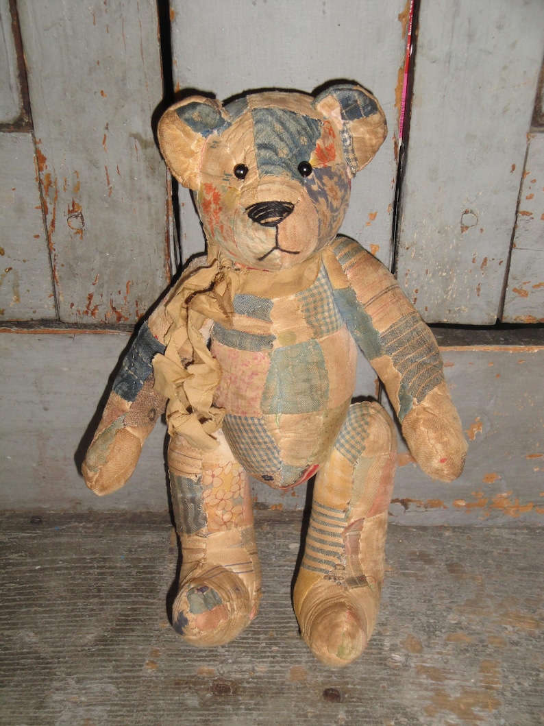 Puede incluir: Un oso de peluche confeccionado con varios parches de tela en tonos apagados de azul, beige y crema. El oso presenta ojos redondos oscuros, una nariz y boca negras bordadas, y una cinta de tela fruncida atada alrededor del cuello.